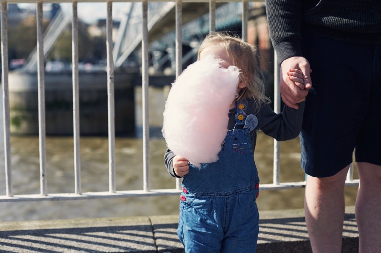 A toddler eats a huge pink candy floss that covers her face next to the River Thames