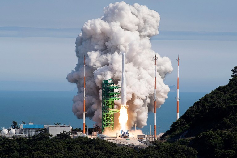 A white rocket begins to take off in front of a green scaffold and a cloud of smoke, with the sea in the distance.