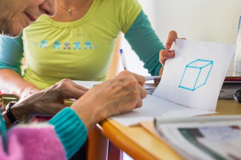 A woman holds a piece of paper with a blue cube-shaped drawing on it next to an elderly woman who is writing on a sheet of paper