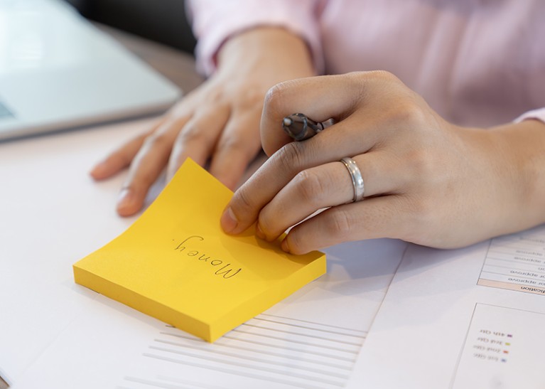 A close-up shot of a young woman's hand writing the word "money" on a yellow sticky note