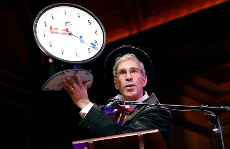 Master of Ceremonies Marc Abrahams holds up the 2016 Ig Nobel award which is a clock with sand timers for hands and letters spelling out Ig Nobel Prize instead of numbers