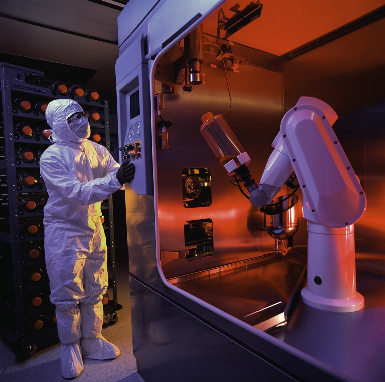 A biotech worker in PPE in a cleanroom controlling a robotic arm.