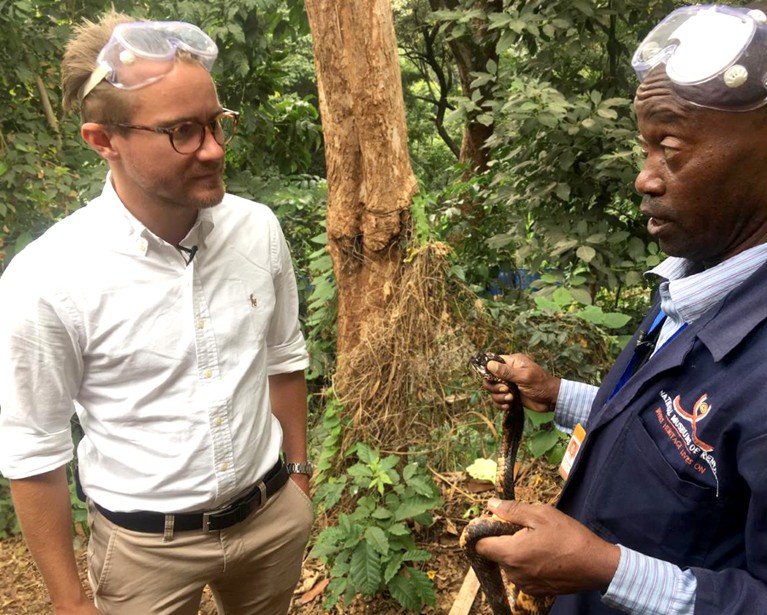 A man in a white shirt with safety goggles on his head listens to a man in a blue jumpsuit who is holding a cobra in a jungle setting