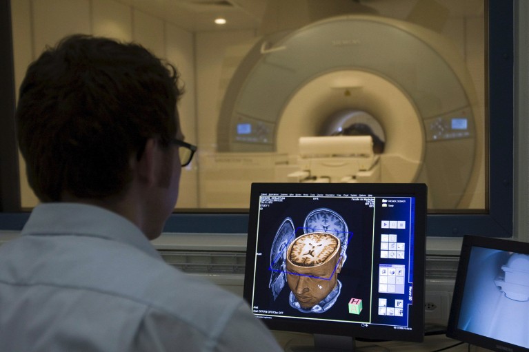 A scientist wearing a lab coat looks at a brain scan on a computer with an MRI machine in the background