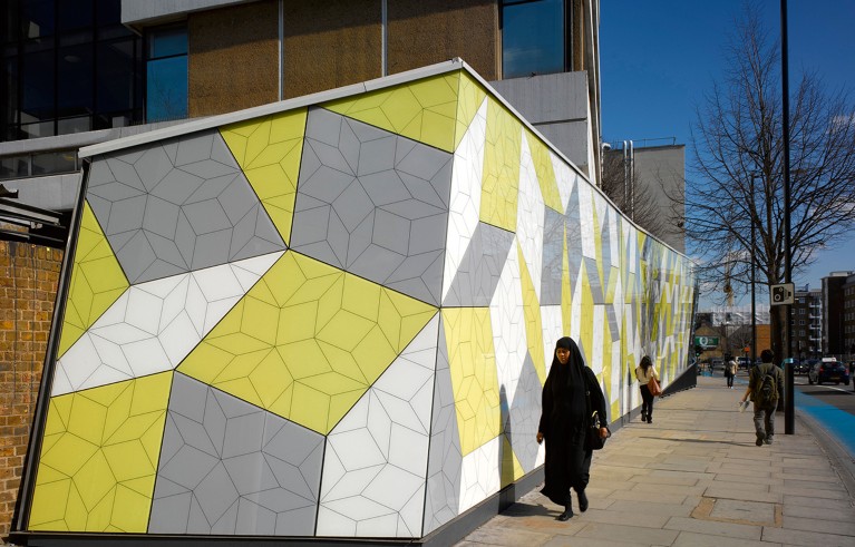 Una mujer vestida con un largo vestido negro pasea junto a una pared blanca, gris y amarilla de un edificio universitario de Londres adornado con diversas formas geométricas relacionadas con los azulejos de Penrose.