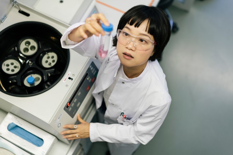 A young scientist in a lab coat and safety glasses examines a centrifuge tube with some pink liquid inside