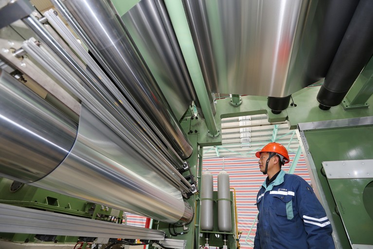 A man in a hard hat and overalls looks at large rollers of thin silver materials overhead