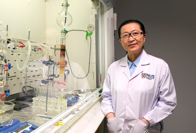 Bin Liu pictured in a lab next to a setup with glass tubes containing coloured liquid substances behind safety glass