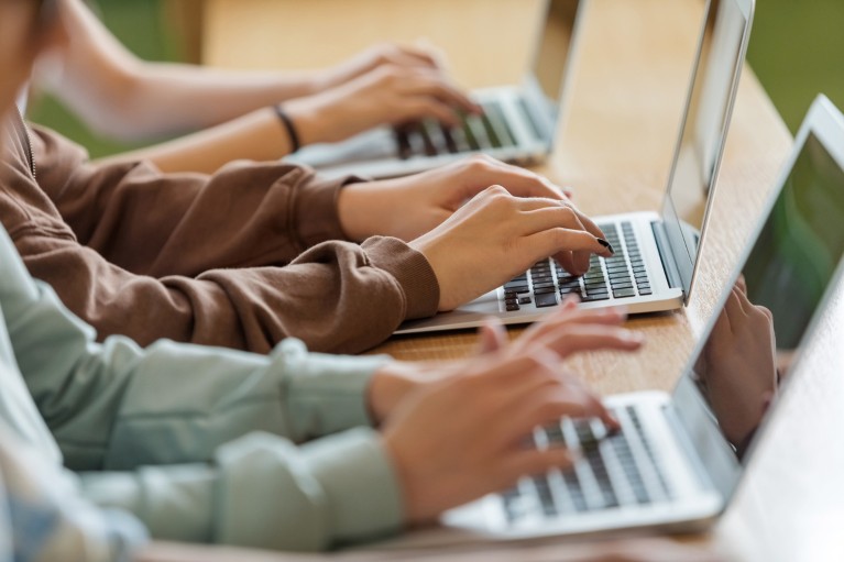 A close-up of the hands of three people typing on laptops at a desk.