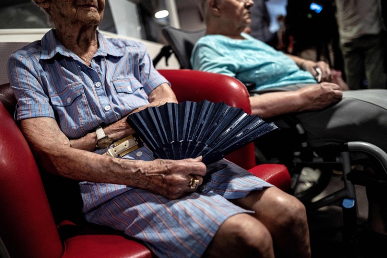 An elderly resident holding a hand fan while sitting in a chair at a care home during a heatwave