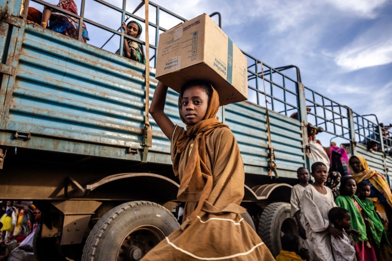 A low-angle view of a young Sudanese girl carrying a cardboard box on her head while making eye contact with the camera