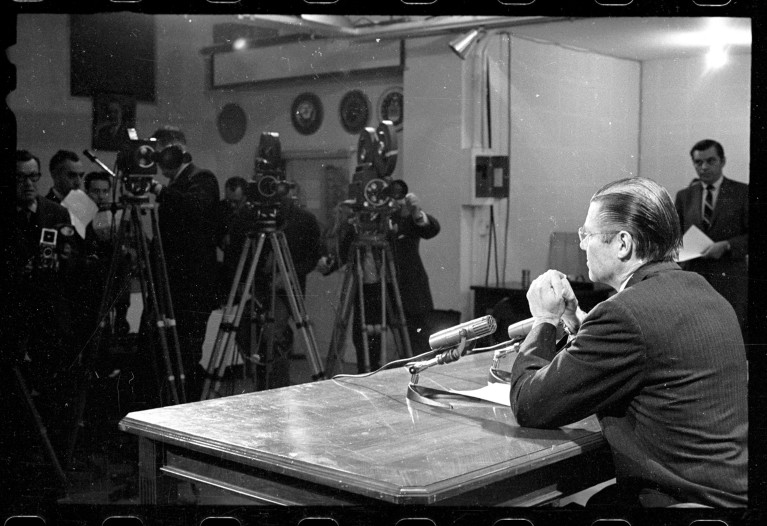 An old black and white film photograph of Robert S. McNamara making an announcement to press