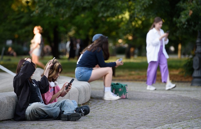 Young women sitting on the street using their smartphones in a park while other women walk in the background, Warsaw, Poland.