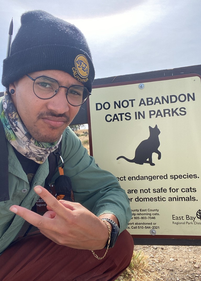 Tyus crouches in front of a warning sign at Oyster Bay Regional Shoreline about abandoning cats in the park