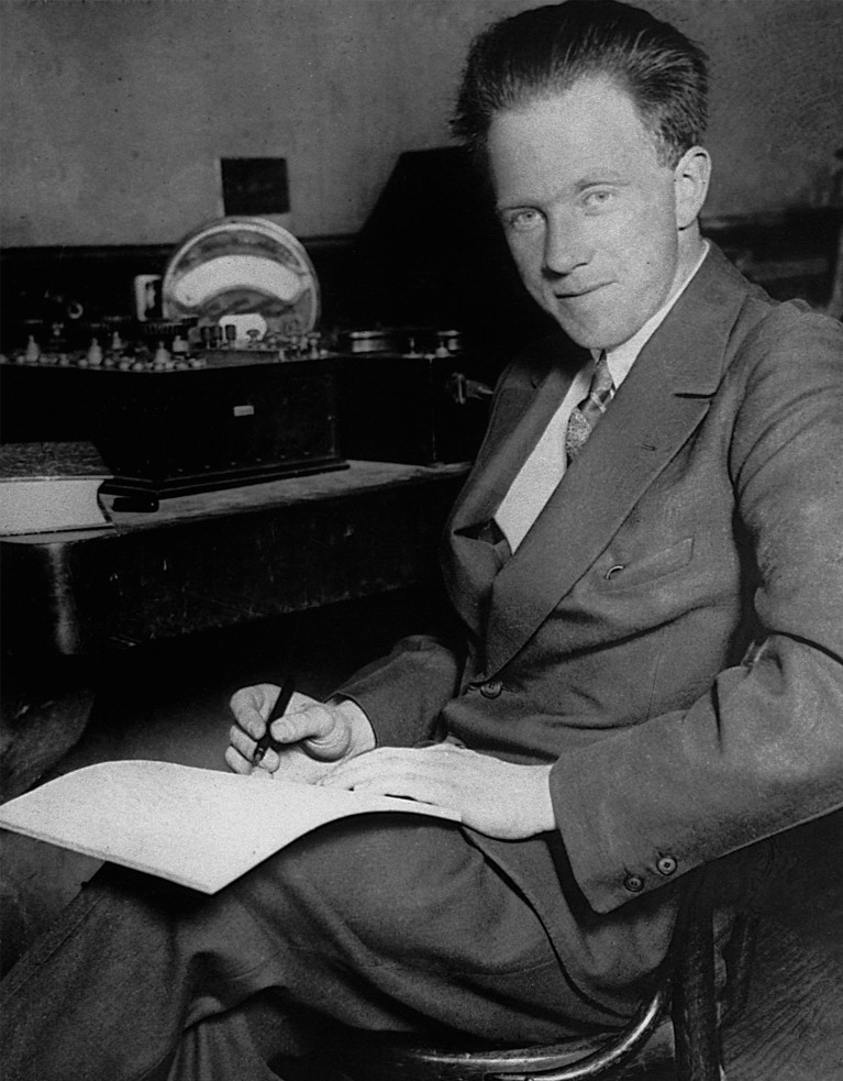 Black and white portrait of Heisenberg sitting on a chair writing on a piece of paper and holding a pen.