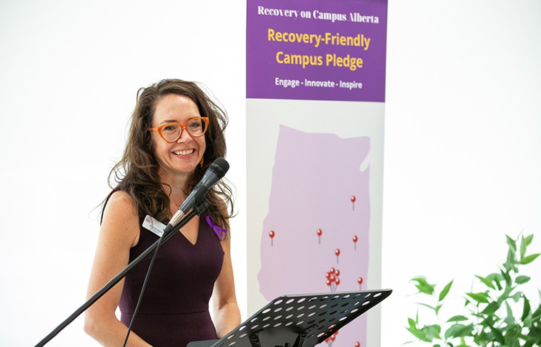 Victoria Burns standing at a podium in front of a poster saying "Recovery-Friendly Campus pledge"