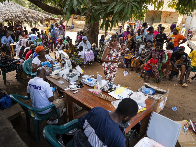 Outside, a woman walks past a table containing laboratory equipment. Three people are sitting at the table, surrounded by a group of people sitting and standing. A large tree at the centre of the image.