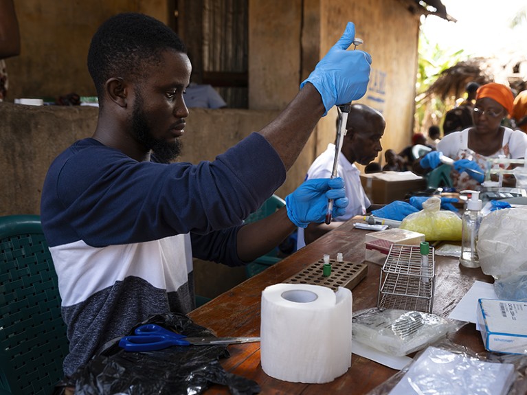 A man sits at a table, holding a pipette and blood sample. Other laboratory equipment is on the table.