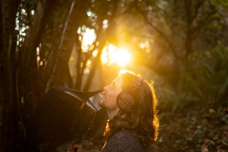 A portrait of Seán Ronayne in a wood with the sun behind him, wearing headphones and holding a large recording device