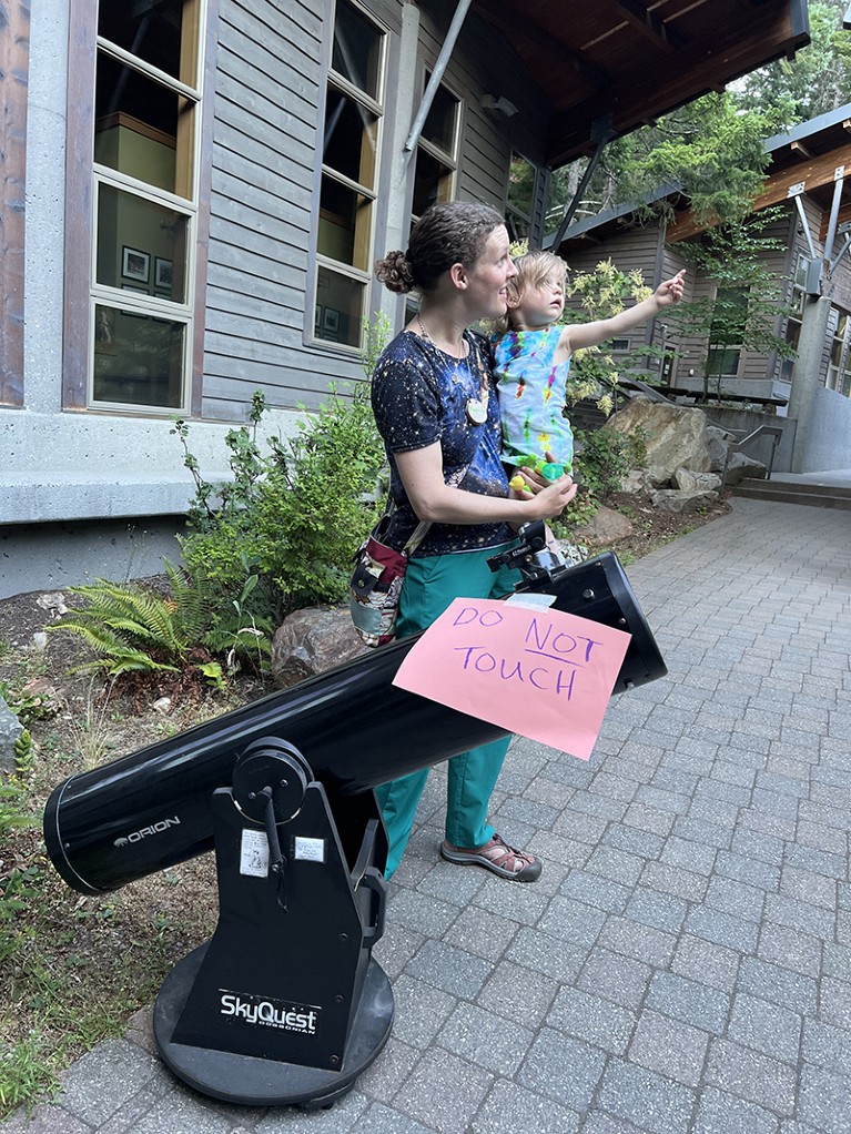 Meredith holding her child standing in front of a telescope