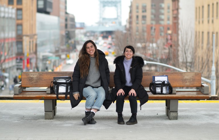 Shai Berman and Tessa Montague sitting on bench at a train station with clear plastic bags and a street with cars and high-rise buildings visible in the background