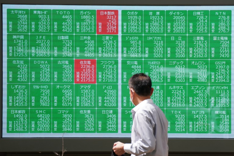 A man wearing a face mask looks at an electronic board displaying stock prices shown in green and red boxes