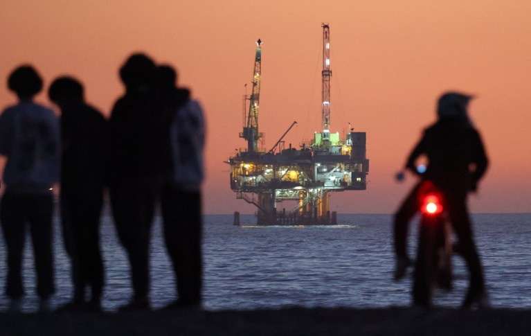 An illuminated oil rig in the sea beyond silhouettes of people gathering on a Californian beach at sunset