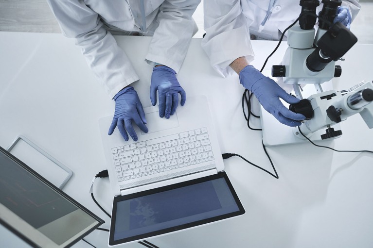 High angle shot of two researchers working in a lab, one types on a laptop while the other uses a microscope.