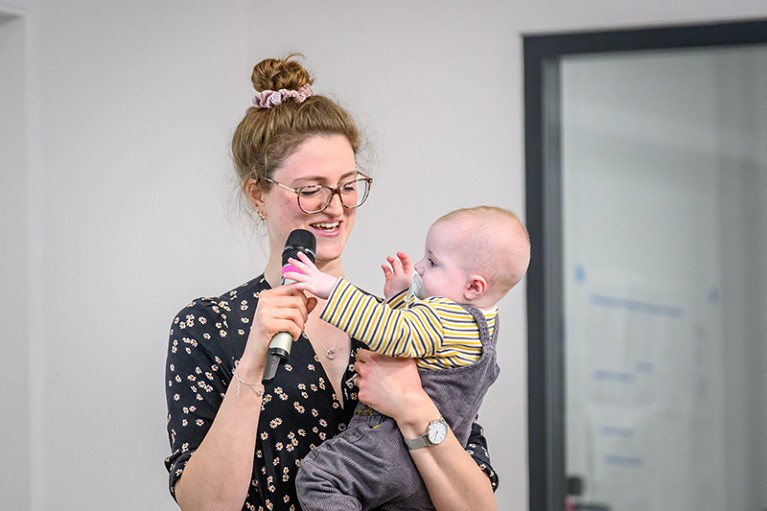 Hannah Chance stands, giving a talk while her son reaches out for the microphone in her hand