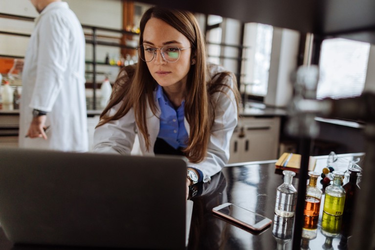 A female scientist wearing a white lab coat typing on a laptop in a laboratory