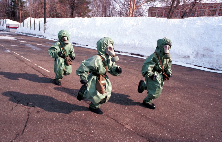 Three small children dressed with gas masks and green plastic protective suits run along the road.