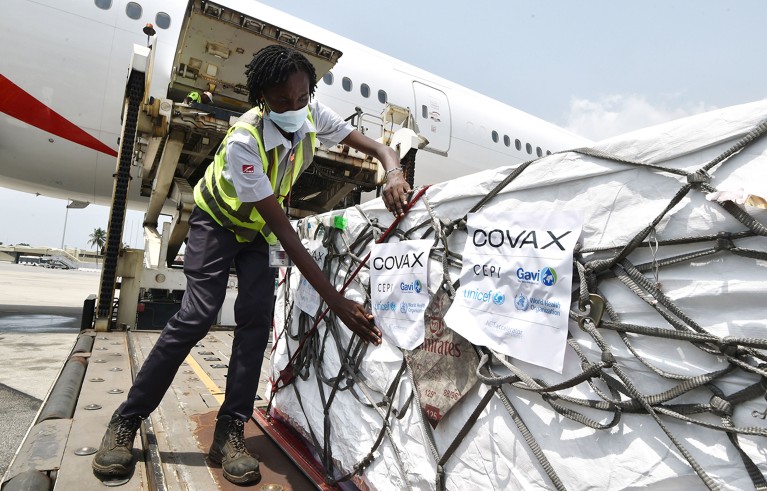 Black lady with face mask and high-vis jacket sorting out a vaccine shipping parcel with cargo airplane behind her.