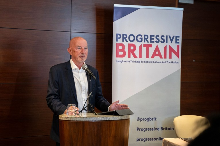 Ewan Kirk speaks at a podium with a banner in the background reading 'Progressive Britain'