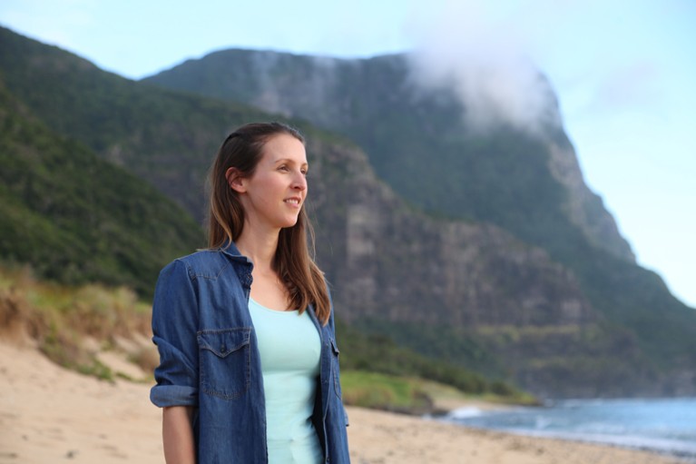 Jennifer Lavers in piedi su una spiaggia guardando il mare con una montagna dietro di lei