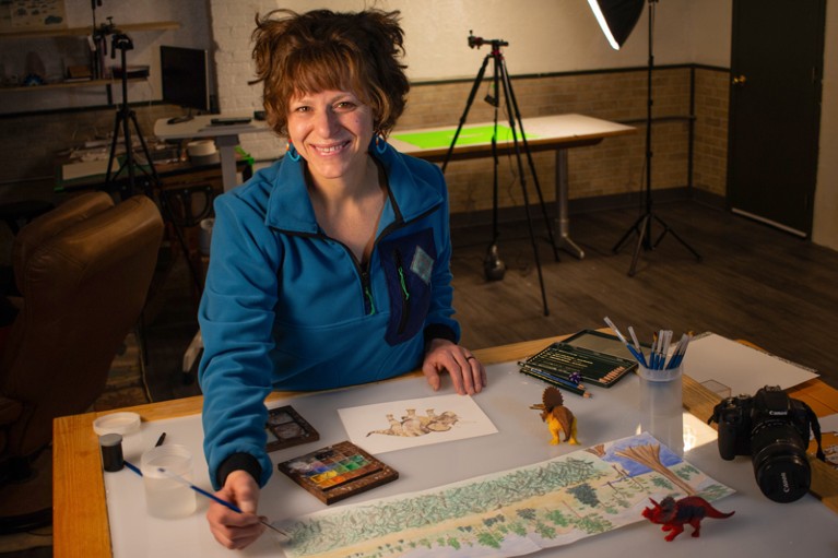 Reilly Dibner sitting at a desk painting with watercolours in an animating studio with lights and tripods in the background