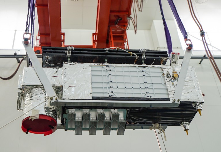 A microsatellite, rectangular in shape and covered in reflective foil, suspended from a crane