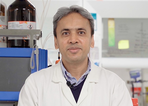 Asian male in white lab coat standing in front of scientific equipment with eyes to camera.