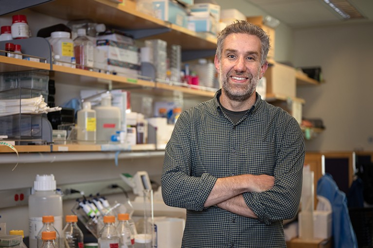 White male with beard and wearing checked shirt. He is standing in front of laboratory shelves with eyes to camera and arms folded.