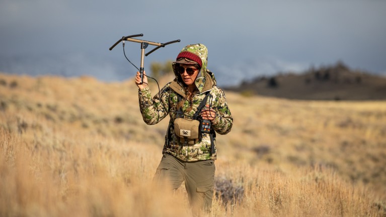 Anna Ortega wearing a camouflage coat and holding an aerial, walking through a field of tall grass while tracking deer in Wyoming