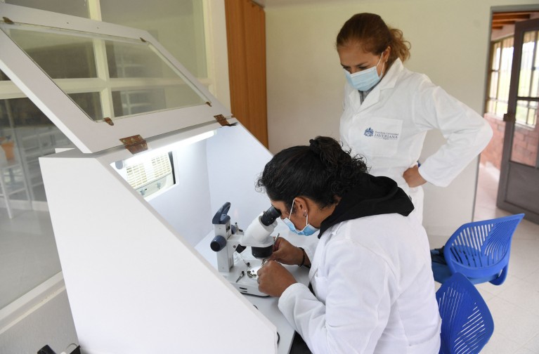 A young female researcher looks down a microscope while another female researcher watches over her in a lab