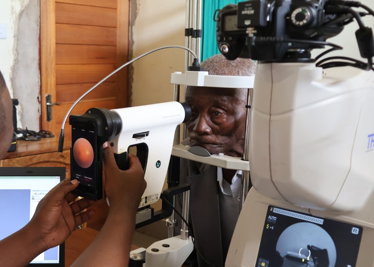 An elderly patient sits for an eye examination, and the picture of his retina is seen on a mobile device held by a doctor