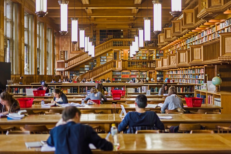 People working and reading at rows of desks in a large wood panelled library with large windows and multiple floors of book shelves