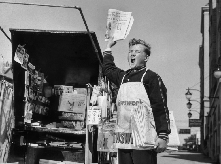 Black and white photo from the 1940s of a newsboy shouting beside a newsstand while holding a newspaper in the air and a bundle of newspapers under his arm