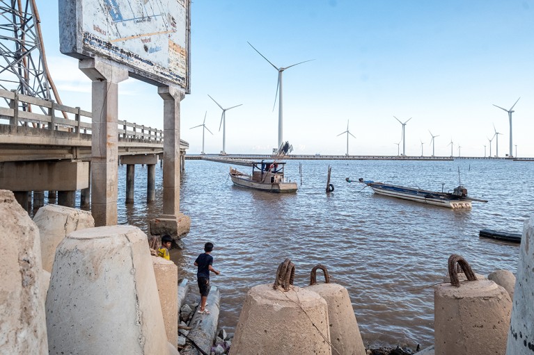 Wind turbines at the Bac Lieu Offshore Wind Farm in Vietnam, with a dock and small boat in the water.