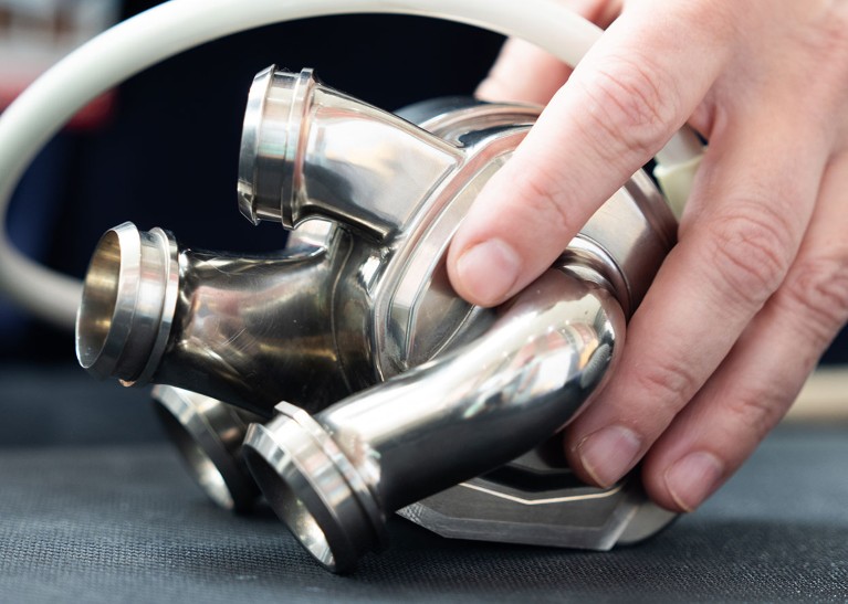 A hand positions a silver metal artificial heart on a dark table surface.