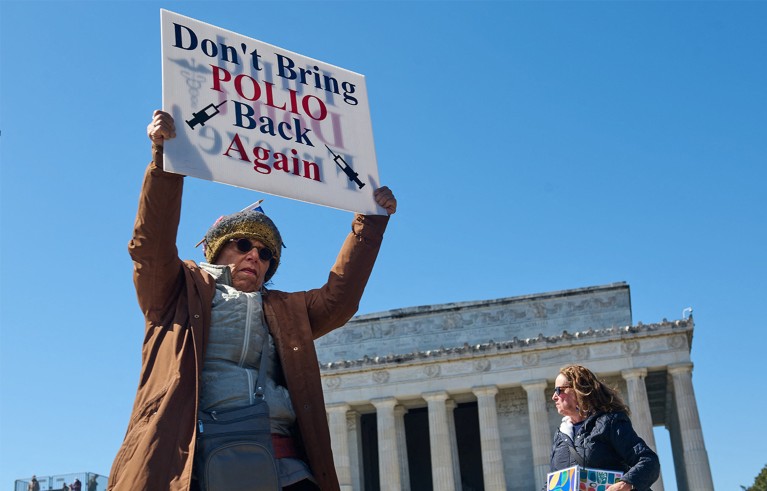 Old person with hat and glasses holds a placard saying Don't bring polio back again. Woman on the left and old building with pillars in the background.