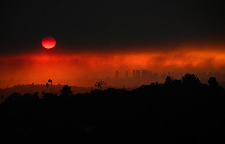 Aerial image shows smoke from wildfires with Los Angeles skyline in the background. Dark thick smokey cloud blocks a red sun.