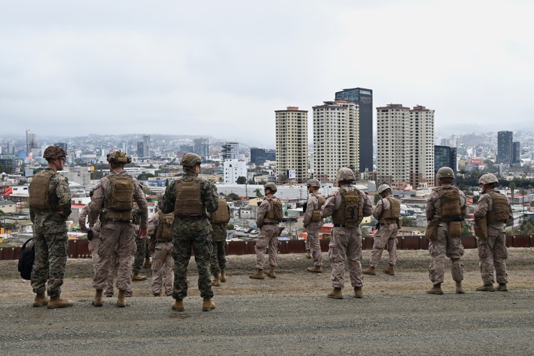 A group of US military personnel in helmets and fatigues look out at a built up area of San Diego at the US-Mexico border