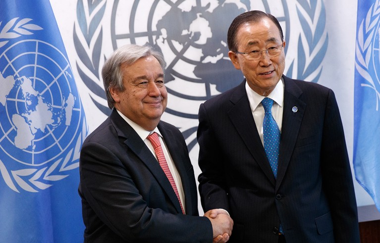 António Gutteres and Ban Ki-Moon shake hands. Logo and blue flag of the United Nations are in the background.