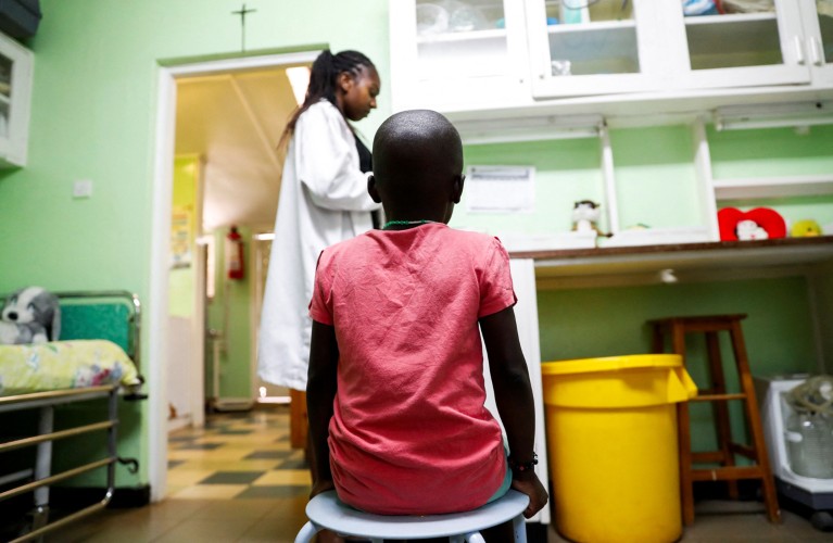 A child sits on a stool with their back to the camera while a nurse wearing a white lab coat prepares to dispense HIV drugs